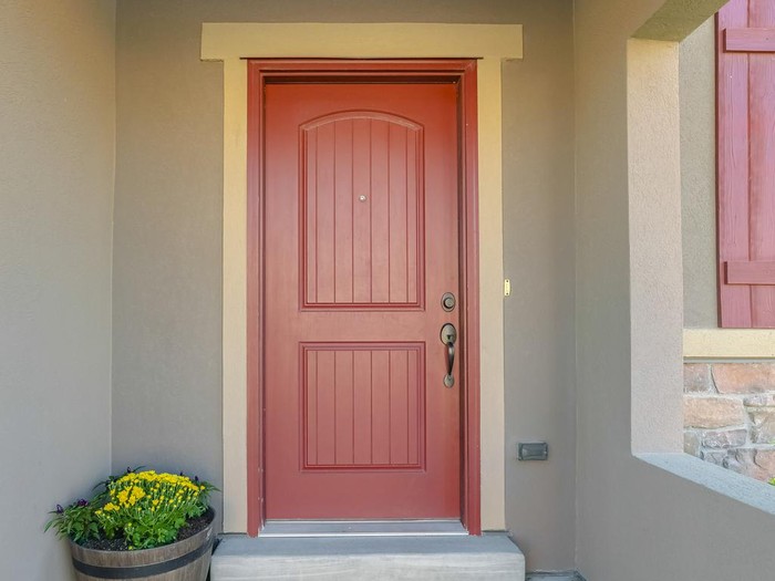 Panorama frame The red front door of a house with concrete exterior wall and shutters on window. Doormat and potted flowers are placed by the doorstep.