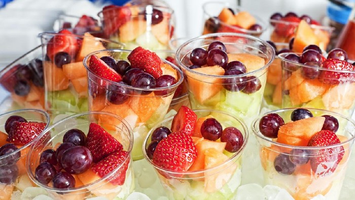 Strawberries, red grapes, cantaloupe offered in plastic cups on ice at a food fair.
