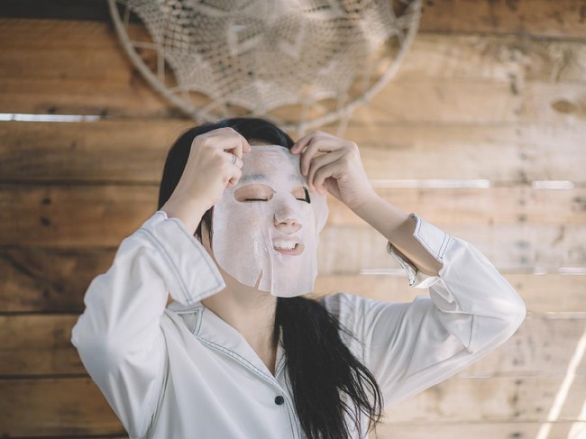 Mature women home in pajama spending cozy morning during isolation period. She wearing sheet mask and have video call, her dog is in bed too