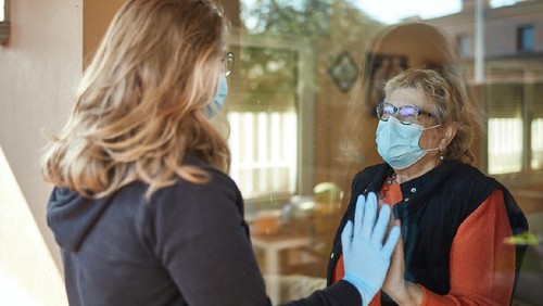 Granddaughter visiting grandmother on her 70s wearing a protective face mask standing indoors at home and touching window with hand  to her granddaughter outdoors in times of COVID-19, granddaughter is wearing mask and gloves.