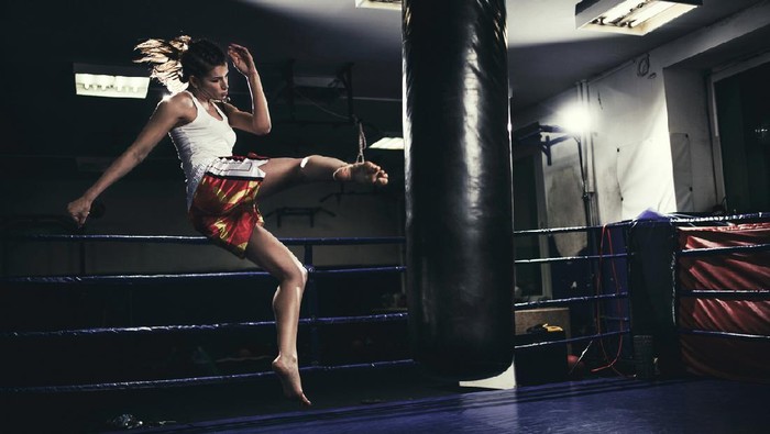Photo of a fit female muay thai fighter training with a punching bag in a boxing hall. She is jumping on a boxing ring in an underground fight club, training kicking.