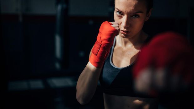 Photo of a fit female muay thai fighter training with a punching bag in a boxing hall. She is jumping on a boxing ring in an underground fight club, training kicking.
