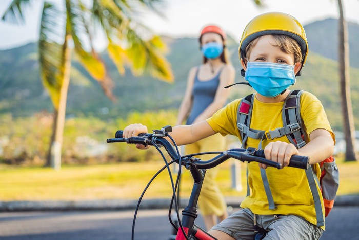 Active school kid boy and his mom in medical mask and safety helmet riding a bike with backpack on sunny day. Happy child biking on way to school. You need to go to school in a mask because of the coronavirus epidemic.