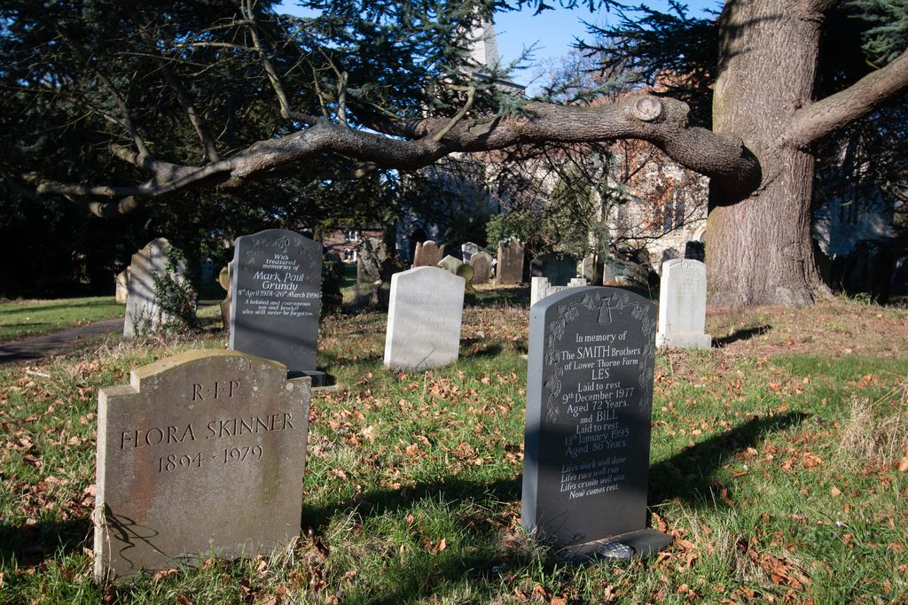 A tombstone for Flora Skinner and the Smith Brothers in a church graveyard. Other names and details are visible on the tombstones. Pluckley has been listed in the Guinness Book of Records for being most haunted village in the UK, with 12 ghosts recorded. The churchyard is haunted by the Red Lady (Lady Dering).