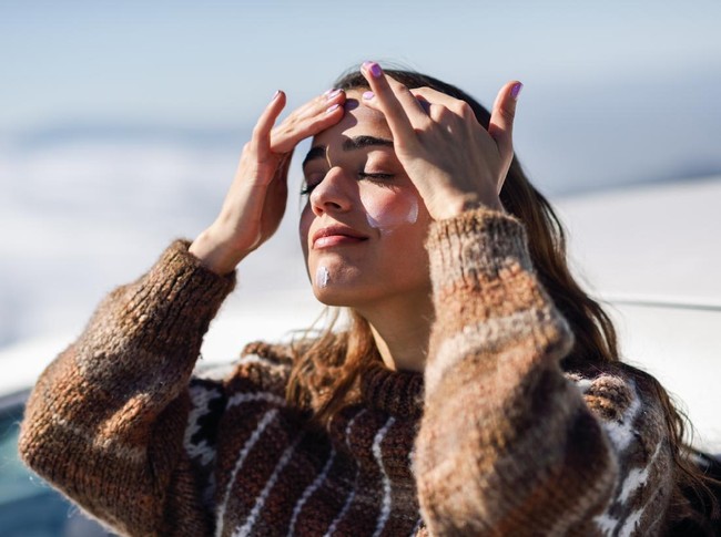 Young woman applying sunscreen on her face in snowy mountains in winter, in Sierra Nevada, Granada, Spain. Female wearing winter clothes.