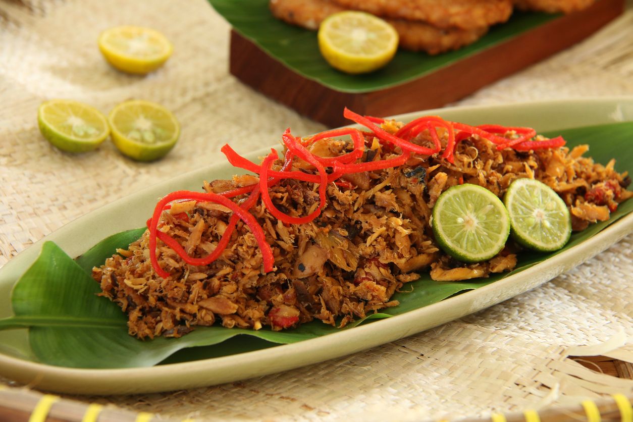 Pampis Ikan Tongkol, a traditional seafood dish from Manado, Sulawesi. Shredded mackerel fish cooked in spicy spice mixture, then garnished with red chili pepper and lime halves. The food is plated on a leaf-shaped ceramic plate that has been lined with a banana leaf of similar size. The plate is arranged on a tray lined with a straw mat. Accompanied with fried tempeh (soybean cakes).