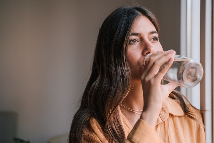 1204691618 young beautiful woman by the window at home drinking water. Lifestyle