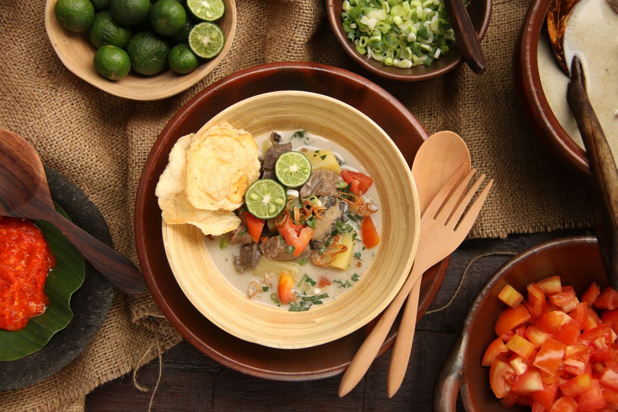Soto Betawi, the popular traditional beef and offal soup from Betawi, Jakarta. The soup dish has been pre-plated in a bamboo bowl with an earthenware plate as the underliner. A pair of wooden spoon and fork is placed on the plate. The soup is surrounded with the broth and condiments.