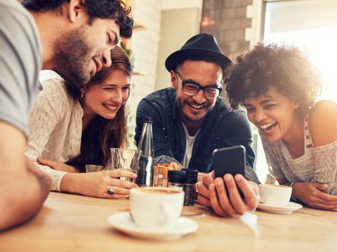 Portrait of cheerful young friends looking at smart phone while sitting in cafe. Mixed race people sitting at a table in restaurant using mobile phone.