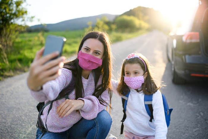 Mother with small daughter taking selfie with smartphone on trip outdoors in nature, wearing face masks.