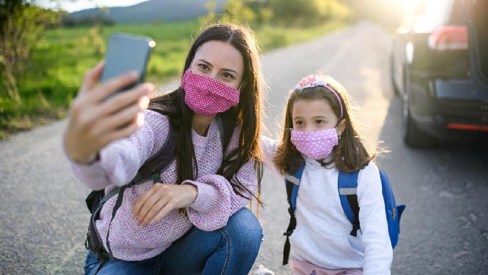 Mother with small daughter taking selfie with smartphone on trip outdoors in nature, wearing face masks.