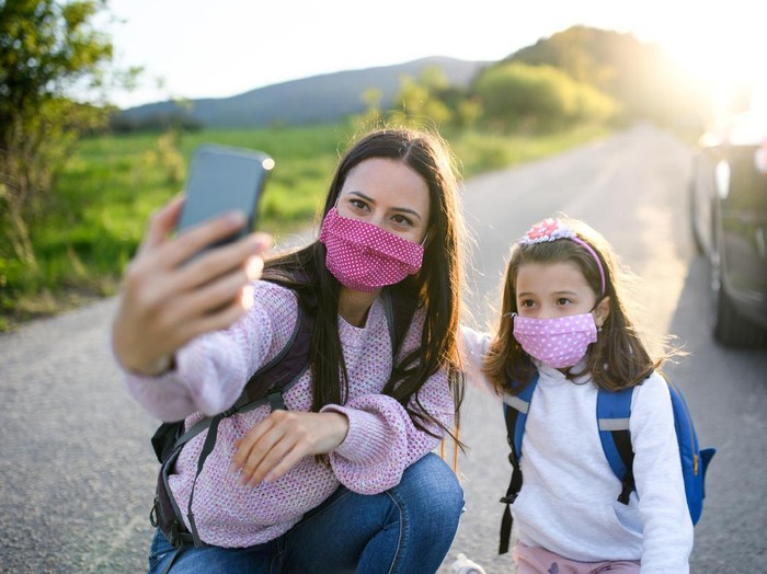 Mother with small daughter taking selfie with smartphone on trip outdoors in nature, wearing face masks.