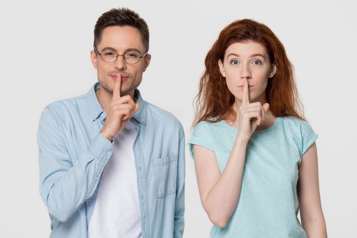 Young mute couple holding fingers on lips mouth to keep it quiet hush secret showing shh shush gesture for conspiracy silence looking at camera isolated on white grey blank studio background