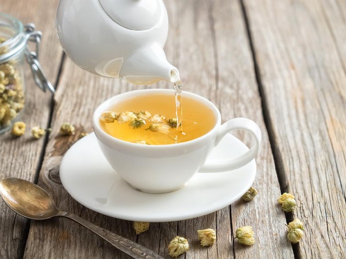 Healthy chamomile tea poured into white cup. Teapot and spoon, glass jar of daisy medicinal herbs.