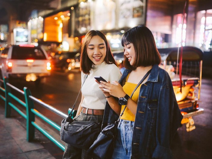 Millenial Vintage toned image of two young, Asian women, walking together, chatting, near Siam station on Bangkok Sukhumvit line. They’re wearing casual street style clothing, enjoying an afternoon together, texting on their cellphones.