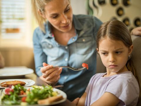 Displeased girl refusing to eat cherry tomato while her mother is feeding her.
