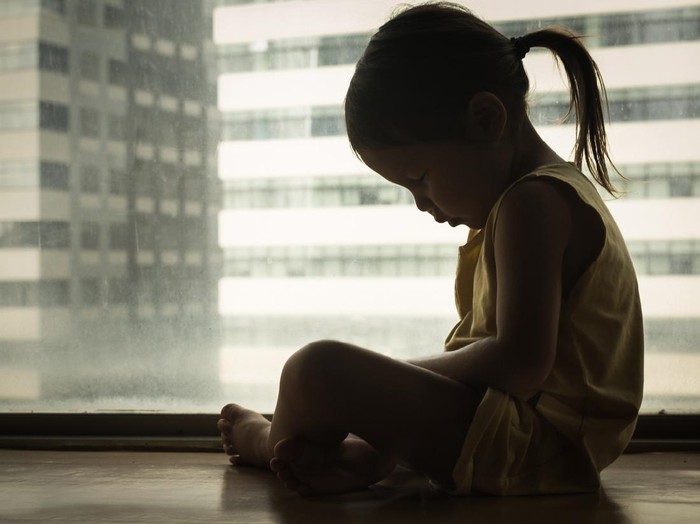 A litte girl sitting next to a window with her head down in sadness. Feeling depressed and hurt.
