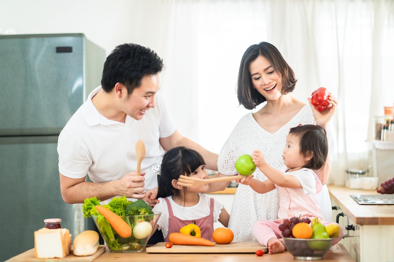 Lovely cute Asian family making food in kitchen at home. Portrait of smiling mother, dad and children standing at cooking counter. kid feeding dad some fruit with smile. Happy family activity together
