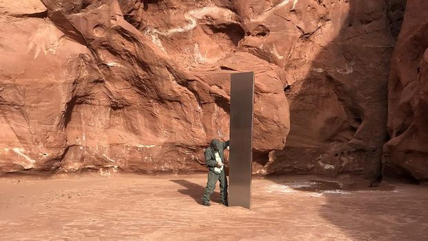 This Nov. 18, 2020 photo provided by the Utah Department of Public Safety shows a Utah state worker inspecting a metal monolith that was found installed in the ground in a remote area of red rock in Utah. The smooth, tall structure was found during a helicopter survey of bighorn sheep in southeastern Utah, officials said Monday. State workers from the Utah Department of Public Safety and Division of Wildlife Resources spotted the gleaming object from the air and landed nearby to check it out. The exact location is so remote that officials are not revealing it publicly, worried that people might get lost or stranded trying to find it and need to be rescued. (Utah Department of Public Safety via AP)