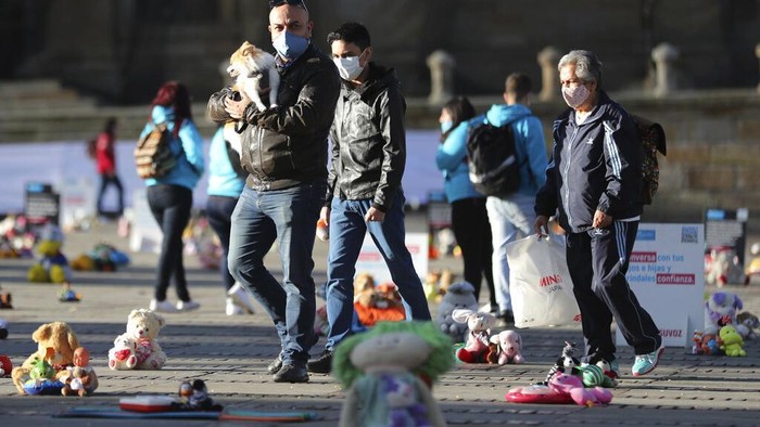People walk past toys representing victims of child violence, that were placed during a protest at Bolivar square in Bogota, Colombia, Monday, Nov. 30, 2020. Hundreds of stuffed animals, dolls and action figures were laid out in Bogota's iconic Bolivar Square to protest violence against children and demand more funding for organizations that help children who have been growing up without their parents. (AP Photo/Fernando Vergara)
