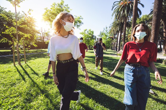 Generation Z teenagers wearing protective face masks and keeping social distancing hanging out together during Covid-19 Coronavirus epidemic spread.