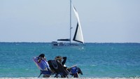 Dua orang wisatawan duduk dengan santai menikmati laut yang dilewati kapal layar di Pantai Miami, Florida. AP Photo/Wilfredo Lee  