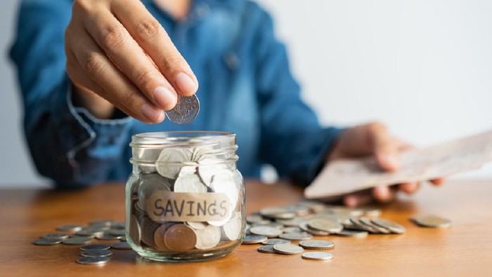 The woman hand is putting a coin in a glass  bottle and a pile of coins on a brown wooden table,Investment business, retirement, finance and saving money for future concept.
