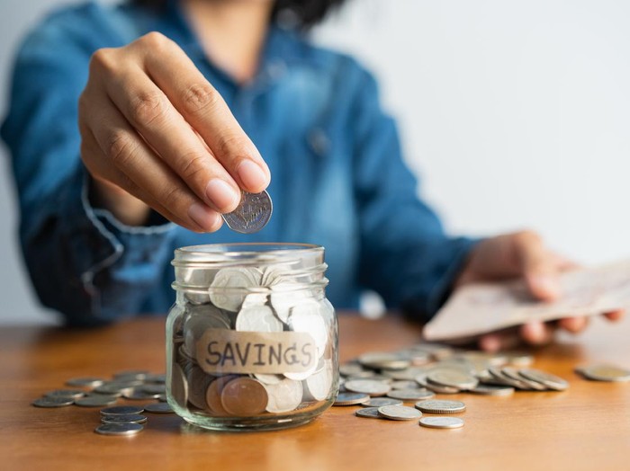 ilustrasi menabung The woman hand is putting a coin in a glass bottle and a pile of coins on a brown wooden table,Investment business, retirement, finance and saving money for future concept.
