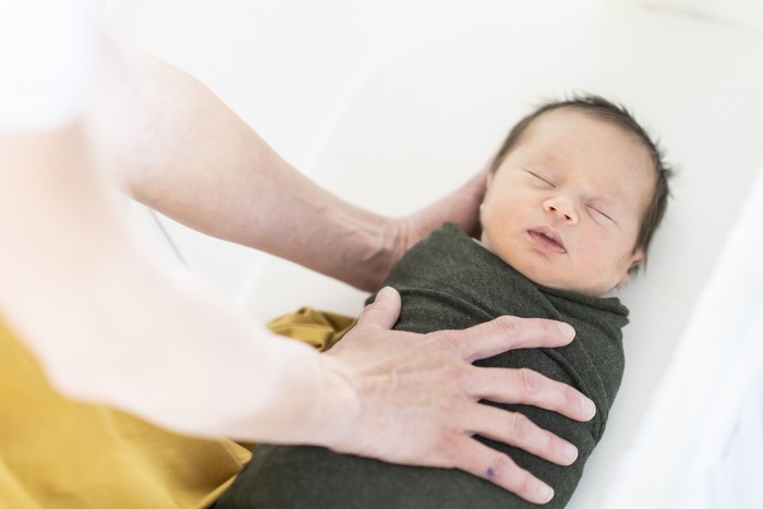 new born Mother gently placing her sleeping newborn baby in the bassinet wrapped tightly in a forest green swaddle.