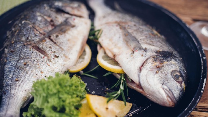 Two whole fish grilled to perfection with a stuffing of lemon slices and rosemary sprigs, presented on a dark plate with lemon and lettuce garnish