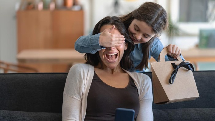 Excited mom being surprised by daughter who is covering her eyes and holding a gift for mothers day - Both smiling