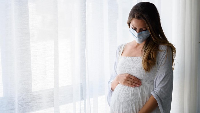 Pregnant woman standing by the big window with face medical mask on. Worries about child birth during pandemic.