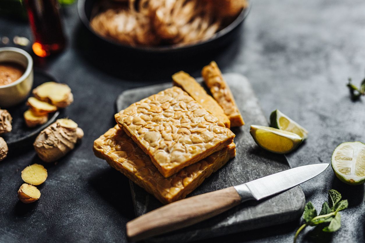 Close-up of tempeh on cutting board with kitchen knife with other ingredients on table. Preparing a vegan dish in kitchen.