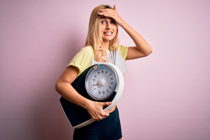 Young beautiful blonde sporty woman on diet holding weight machine over pink background stressed with hand on head, shocked with shame and surprise face, angry and frustrated. Fear and upset for mistake.