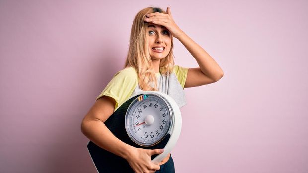 Young beautiful blonde sporty woman on diet holding weight machine over pink background stressed with hand on head, shocked with shame and surprise face, angry and frustrated. Fear and upset for mistake.