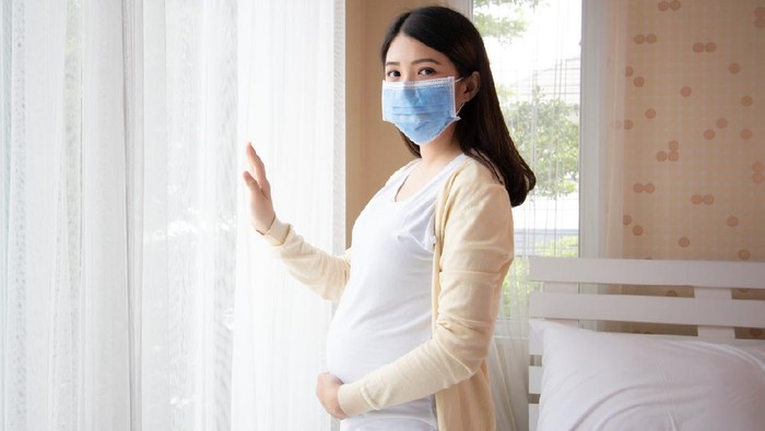 Ilustrasi ibu hamil pakai masker Young beautiful pregnant asian woman standing and touching belly near the window in bedroom. Pregnancy, motherhood wearing virus protection mask mouth concept.