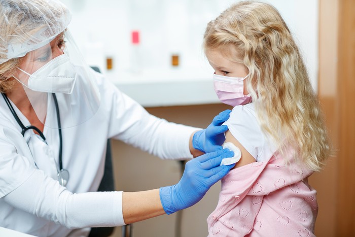 Female doctor in full protective workwear cleaning girls arm before injecting COVID-19 vaccine, girl wearing protective face mask