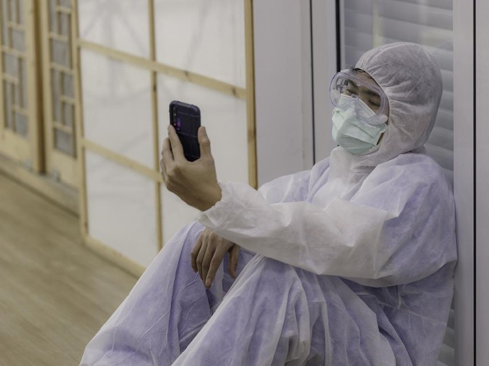 young Asian doctor, dressed in anti-virus clothing, sits on the floor tired and uses a smartphone to make a video call to his family. corona virus concept.