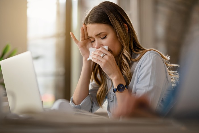 1185448974 Young businesswoman with clpd and flu virus blowing nose while working in the office.