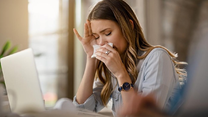 1185448974 Young businesswoman with clpd and flu virus blowing nose while working in the office.