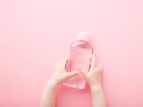 Baby girl little hands holding transparent plastic bottle of oil on light pink table background. Pastel color. Care about soft body skin. Closeup. Point of view shot. Top down view.