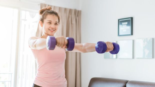 aktivitas olahraga saat pandemi A woman wearing sports clothes raised a dumbbell exercise in the living room. She is training at home. woman in sportswear with Purple dumbbells in her hands.