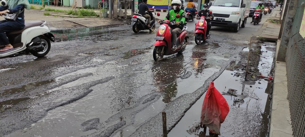 Jalan Caman Raya rusak berlubang hingga digenangi air selokan.