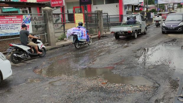 Jalan Caman Raya rusak berlubang hingga digenangi air selokan.