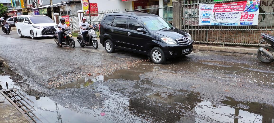 Jalan Caman Raya rusak berlubang hingga digenangi air selokan.