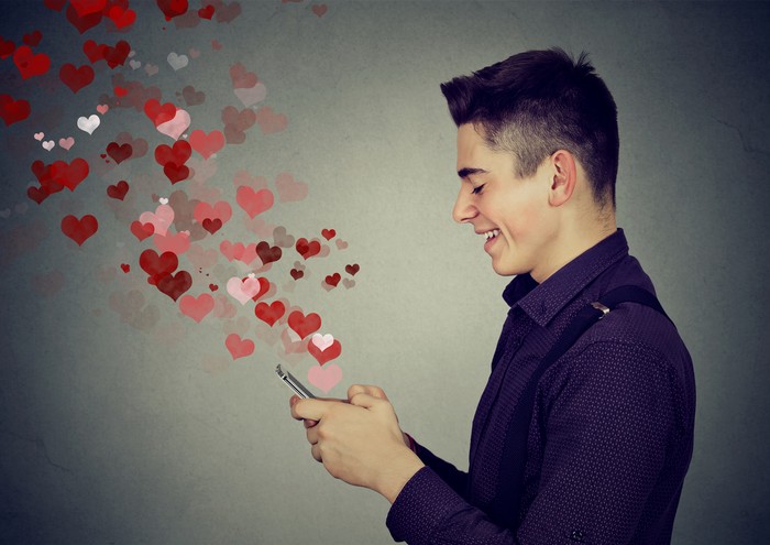 Side profile happy man sending love sms text message on mobile phone with red hearts flying away from screen isolated on gray wall background. Positive human emotions