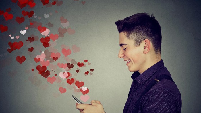 Side profile happy man sending love sms text message on mobile phone with red hearts flying away from screen isolated on gray wall background. Positive human emotions