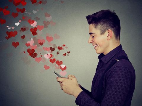 Side profile happy man sending love sms text message on mobile phone with red hearts flying away from screen isolated on gray wall background. Positive human emotions