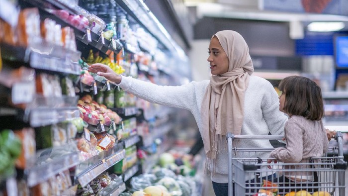 A young Muslim mother and daughter walk the produce aisles of the grocery store together shopping for groceries.  They are both dressed casually and the mother is wearing a Hijab.  The mother is reaching for products on the shelf while pushing a shopping cart with her daughter seated in it.