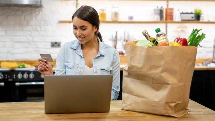 Happy woman at home buying groceries online on her laptop computer and paying by credit card